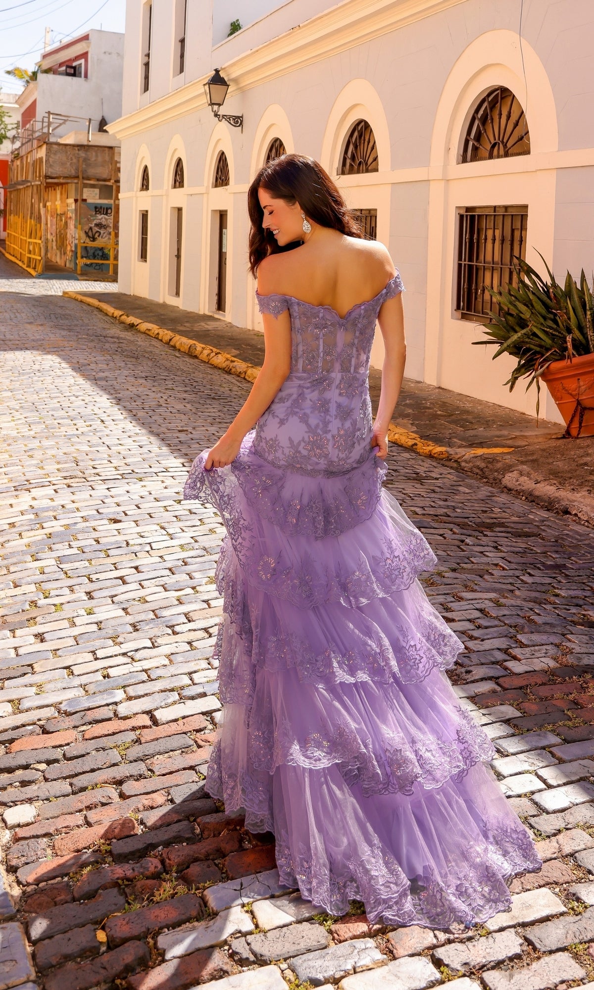 A woman in the Off-Shoulder Long Lace Ruffled Prom Dress R1301 stands on a sunlit cobblestone street lined with white buildings and arched windows.